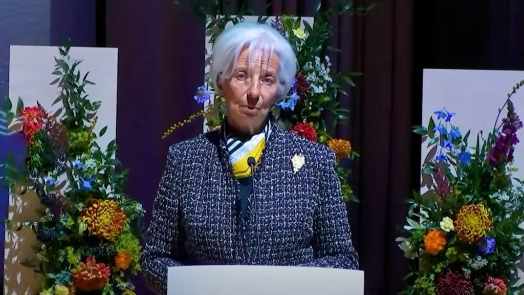 Lagarde with short white hair stands at a podium speaking into a microphone, wearing a dark tweed jacket, a yellow scarf and a brooch, flanked by tall colorful floral arrangements.