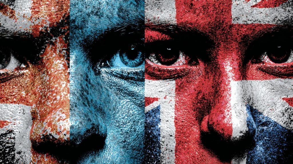 Close-up of a face divided into vertical panels painted with textured red, white and blue flag motifs (including Union Jack elements), emphasizing intense eyes and cracked, weathered skin.