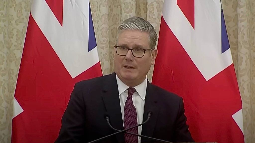Keir Starmer with gray hair and glasses wearing a dark suit and patterned purple tie speaking at a podium, flanked by two large British flags and a beige patterned curtain backdrop.