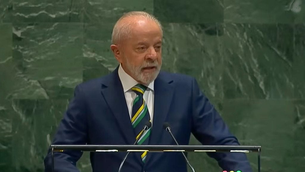 Lula with a short white beard and balding head in a navy suit and green-and-yellow striped tie speaking at a podium against a dark green marble wall.