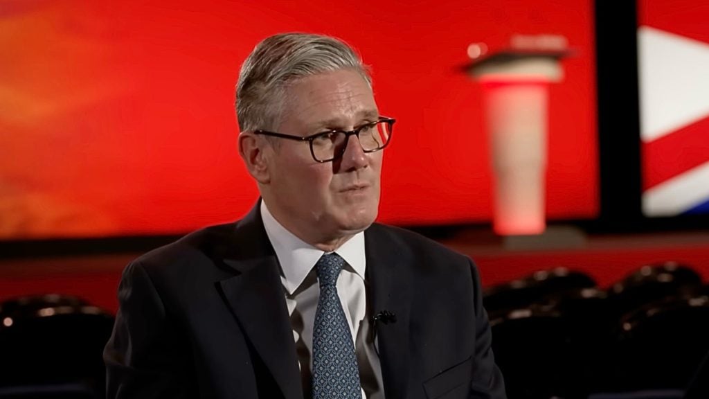 Starmer with gray hair and glasses wearing a dark suit and patterned blue tie, seated and speaking in a dim auditorium in front of a bright red stage backdrop with a blurred podium and part of a red-white-blue flag,