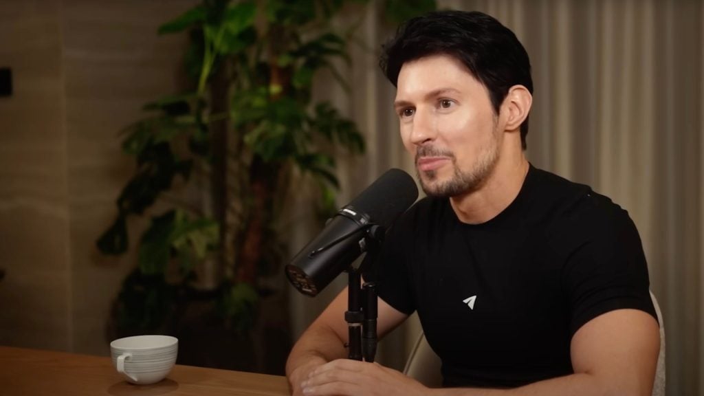 Durov with short dark hair and a trimmed beard speaking into a studio microphone at a wooden table, wearing a black t-shirt with a small logo; a white cup sits on the table and a potted plant and curtain are visible in the background.