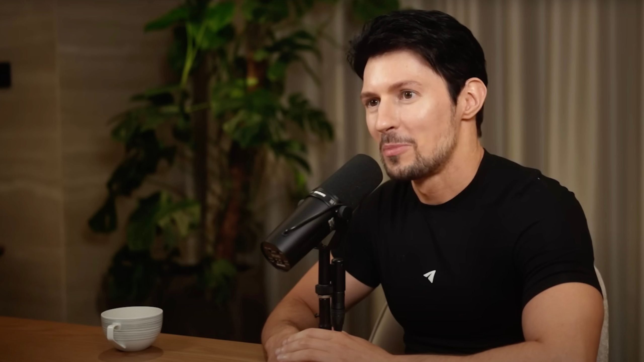Durov with short dark hair and a trimmed beard speaking into a studio microphone at a wooden table, wearing a black t-shirt with a small logo; a white cup sits on the table and a potted plant and curtain are visible in the background.