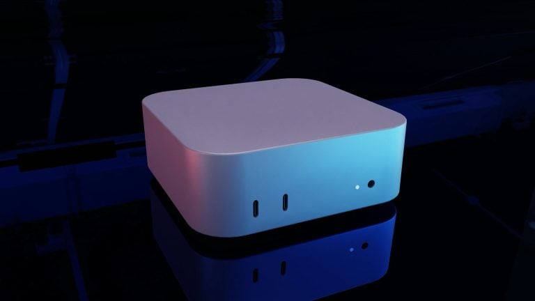 Compact silver rounded-square desktop device with two vertical USB-C ports and a small circular indicator on the front, bathed in blue and purple lighting and resting on a glossy reflective surface