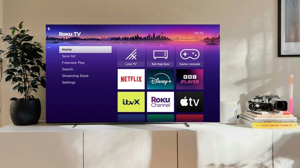 Flat-screen TV on a white media console showing the Roku TV home screen with a purple skyline background and app tiles (Netflix, Disney+, BBC iPlayer, ITVX, Roku Channel, Apple TV), a potted plant to the left and a stack of books with a camera and candle to the right.