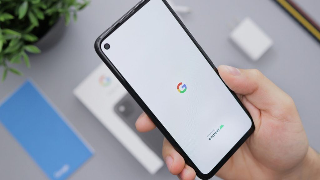 Hand holding a black-cased smartphone displaying a white startup screen with the multicolored Google logo and 'Powered by Android' text, with a blurred phone box, charger and plant in the background.
