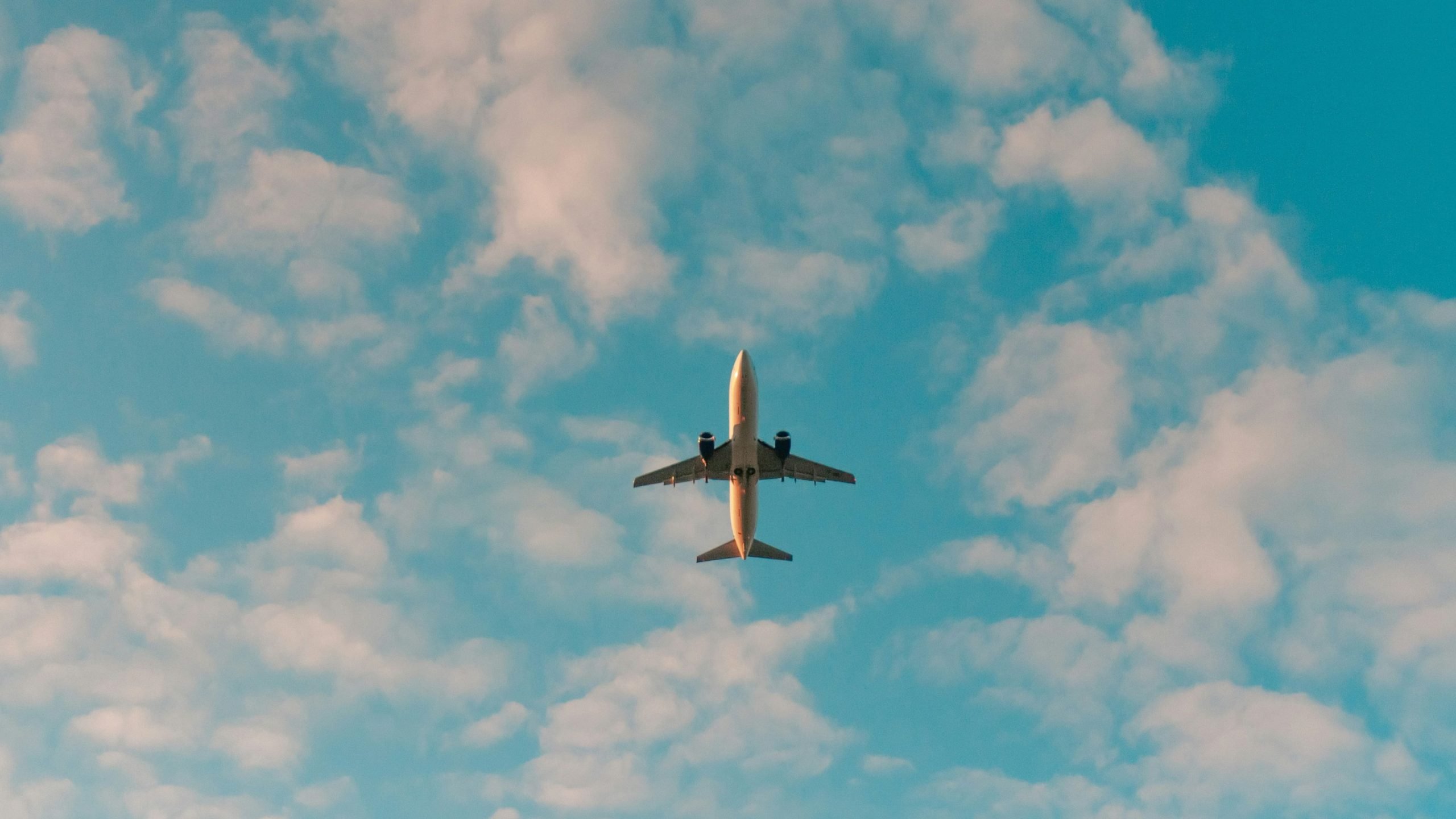 Twin-engine passenger airplane viewed from below, centered against a bright blue sky scattered with soft white clouds.