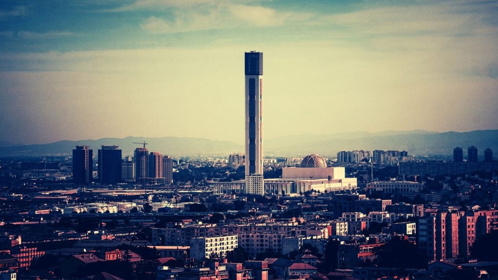 A tall, slender modern tower dominates the center of a sprawling cityscape, with a large domed white building at its base, surrounding mid-rise buildings in the foreground and hazy hills under a pale blue sky in the background.