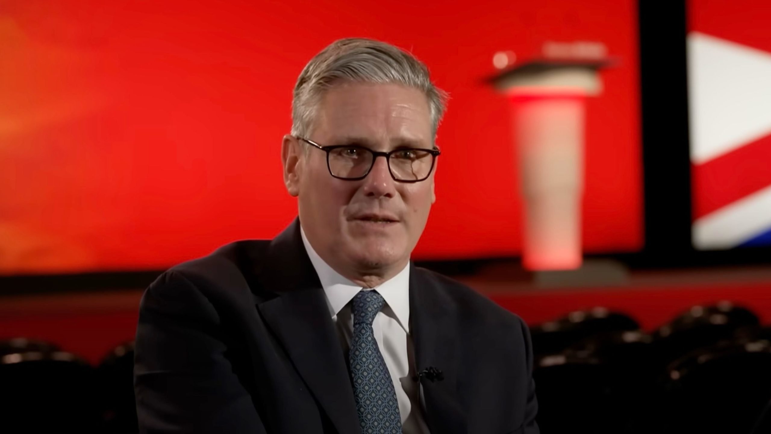 Starmer with gray hair and glasses wearing a dark suit and patterned tie, seated in front of a red stage background with a blurred lectern and a red-white-blue flag element.