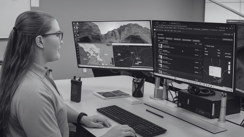 Person with glasses and long hair tied back sits at a desk typing on a keyboard while looking at two large monitors showing maps and a dark application, with a radio, tablet, pen cup and headphones on the desk.