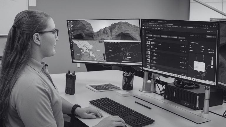 Person with glasses and long hair tied back sits at a desk typing on a keyboard while looking at two large monitors showing maps and a dark application, with a radio, tablet, pen cup and headphones on the desk.