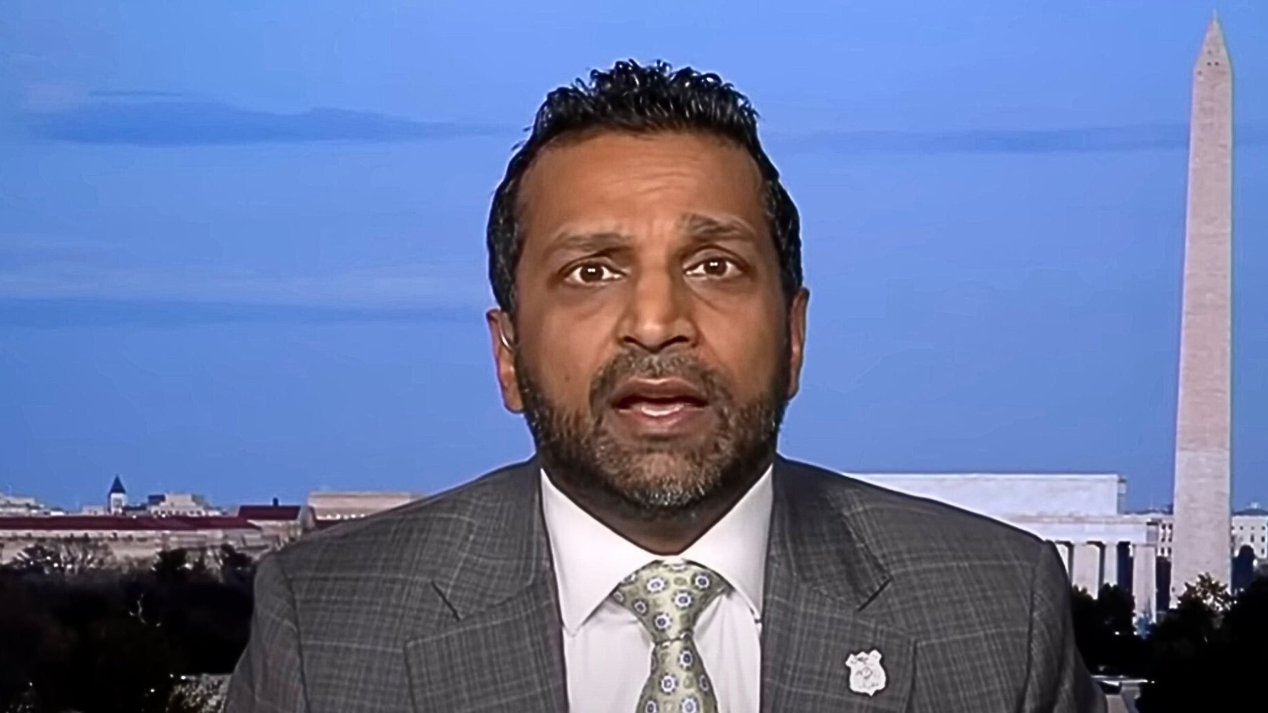 Close-up of a bearded man in a gray checked suit, white shirt and patterned tie speaking on camera, with the Washington Monument and the Lincoln Memorial visible against a blue sky in the background.