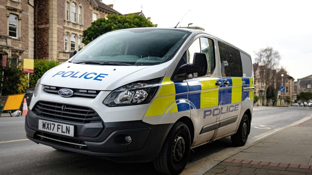 White Ford police van with blue-and-yellow markings and POLICE lettering parked on a city street in front of stone buildings.