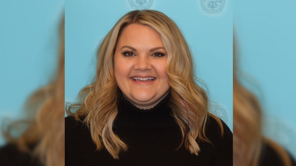Cochran with shoulder-length wavy blonde hair and light makeup, wearing a black turtleneck sweater against a light blue backdrop with faint circular emblems.