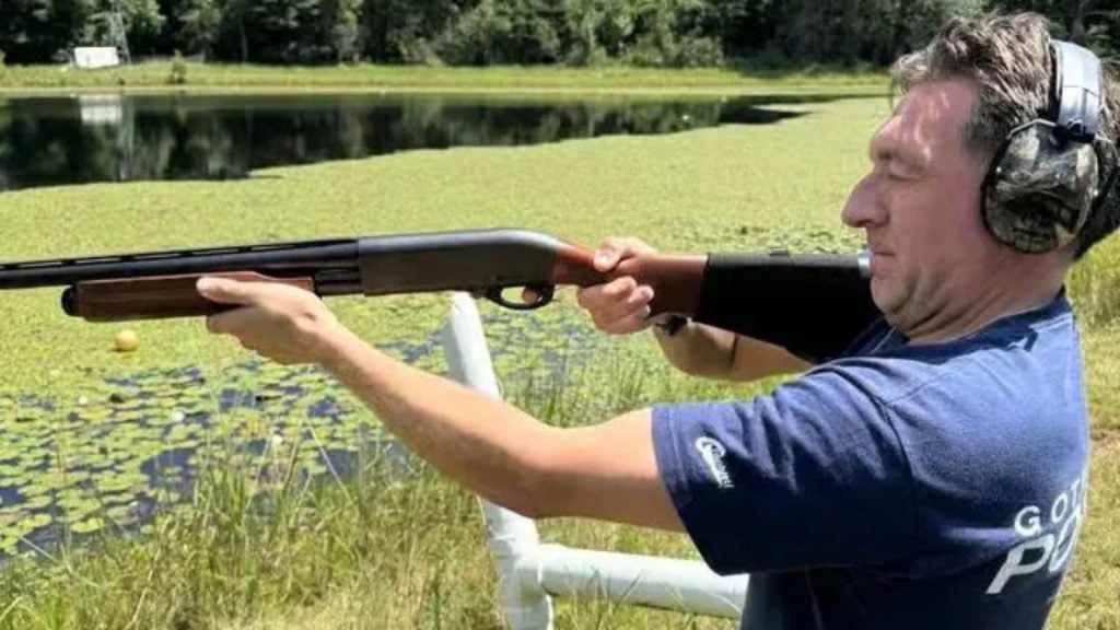 Man in a blue shirt and hearing protection aims a pump-action shotgun across a lily-covered pond while another person’s arm supports the gun near the stock.