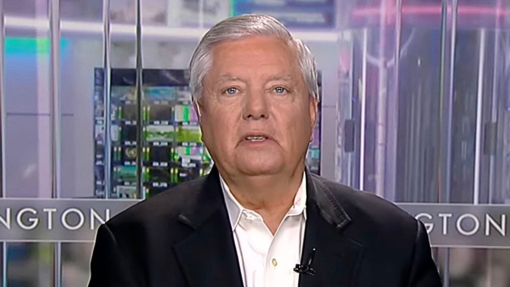 Older man with short gray hair wearing a dark blazer and white shirt speaking on camera in a studio with glass panels and blurred monitors behind him.