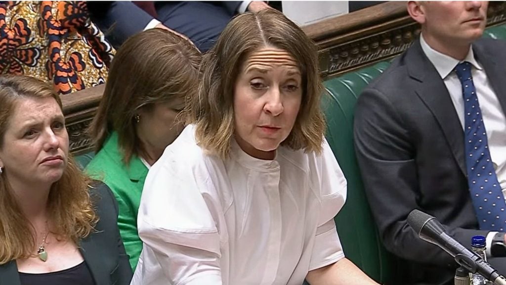 Kendall with shoulder-length brown hair wearing a white blouse seated on a green leather bench in a formal parliamentary chamber, flanked by other seated people and a microphone visible in the foreground.