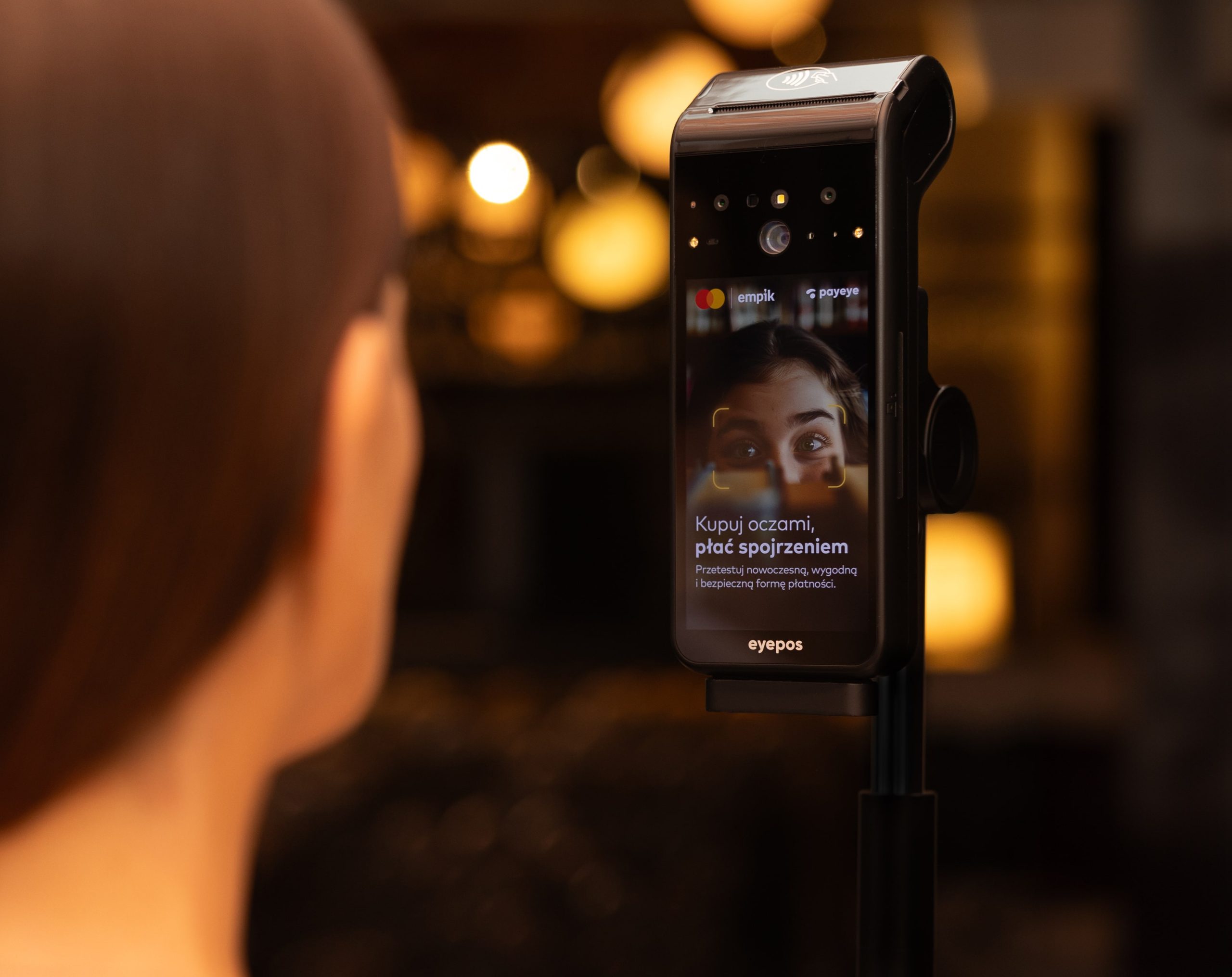 Detailný záber platobného terminálu eyepos s rozpoznávaním očí a tváre, ktorý na obrazovke zobrazuje ženské oči a poľský výzvu „Kupuj oczami, płać spojrzeniem“ s rozmazaným zákazníkom v popredí a teplými bokeh svetlami v pozadí.