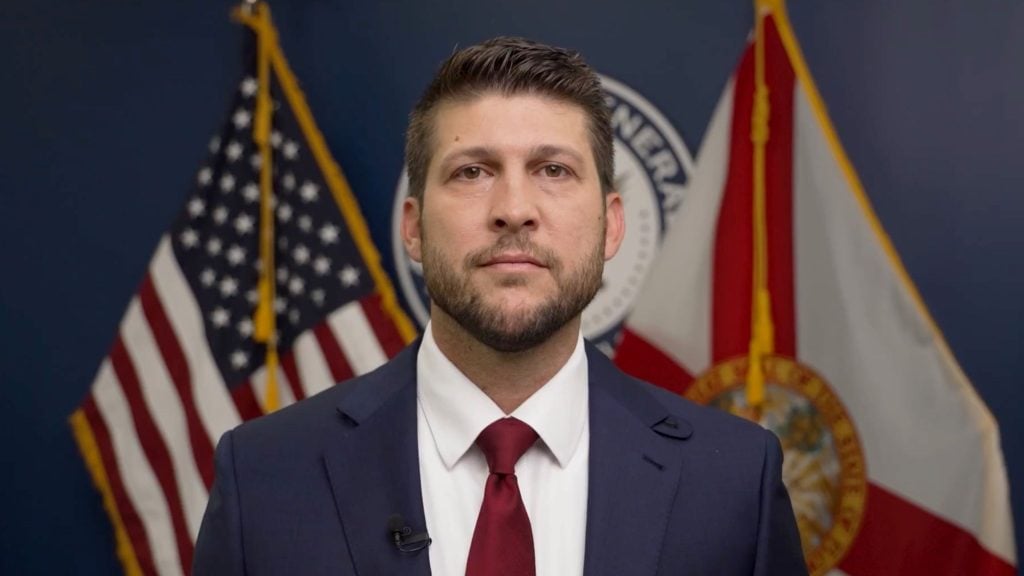 Florida’s Attorney General James Uthmeier in a dark blue suit, white shirt and burgundy tie facing the camera with U.S. and Florida state flags and a circular official seal on a dark blue wall behind him.
