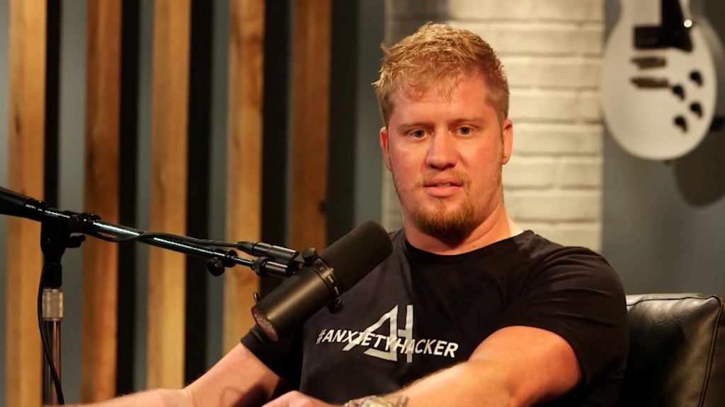 Krottinger with short hair and a trimmed beard seated in a podcast studio speaking into a microphone, wearing a black t-shirt that reads '#ANXIETYHACKER,' with wooden slats, a white brick wall and a white electric guitar visible behind him.