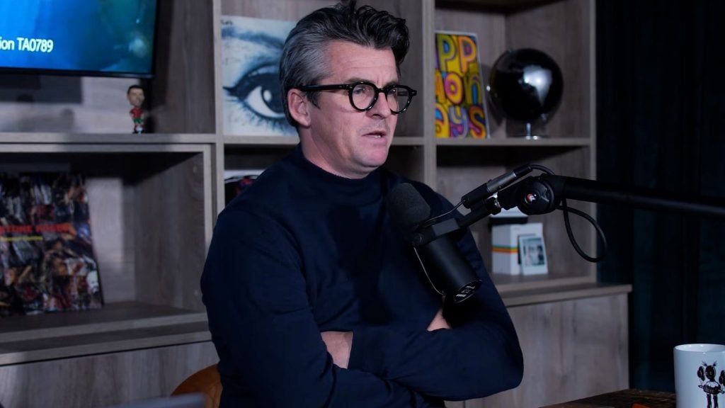 Barton with round black glasses sits with arms crossed speaking into a boom microphone in a podcast studio, with wooden shelving behind holding an eye illustration, a colorful pop-art print, a small globe and a white mug on the table.