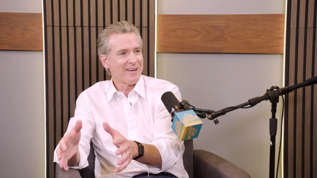Newsom with slicked-back gray hair wearing a white button-down shirt, gesturing with both hands while speaking into a microphone with a blue foam flag in a studio with vertical wood slats and a wooden panel backdrop.