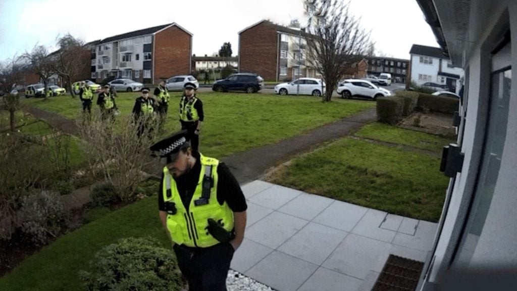 A line of police officers in high-visibility yellow vests approach a suburban house along a paved path across a grassy front lawn, with police vehicles and brick apartment buildings visible in the background.