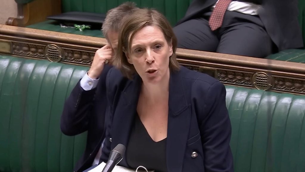 Phillips in a dark blazer speaking with her mouth open into a microphone while seated on green leather benches in a parliamentary chamber, with papers and another person partially visible behind her.