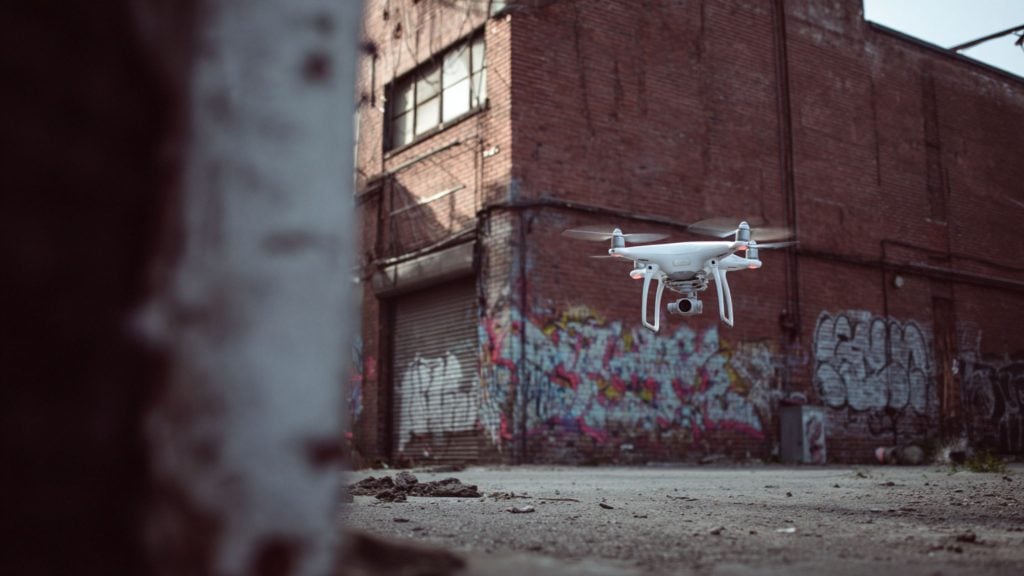 White quadcopter drone with a visible camera and spinning propellers hovering over a cracked concrete lot in front of a graffiti-covered red brick industrial building, with a blurred pillar in the left foreground.