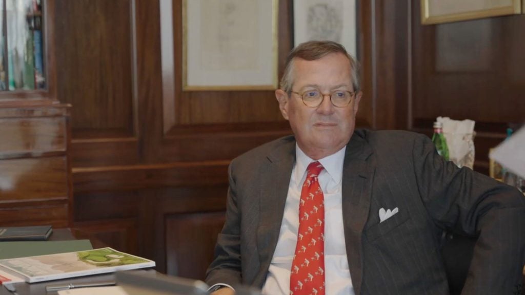 Stephens wearing eyeglasses, a dark pinstripe suit and a red tie with a small white pattern sits back in a chair in a wood-paneled office, looking slightly to the right with papers, books and a bottle of water on the desk beside him.