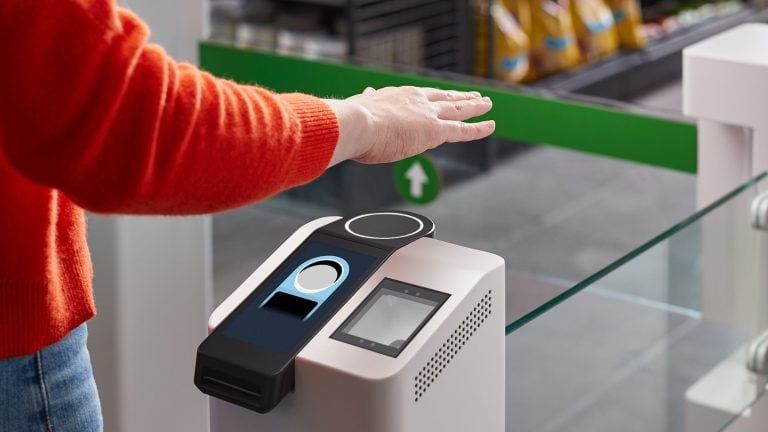 A person wearing a red sweater holds their hand palm-down over a contactless payment/access scanner on a white kiosk at a store entrance, with a green directional barrier and product shelves in the background.