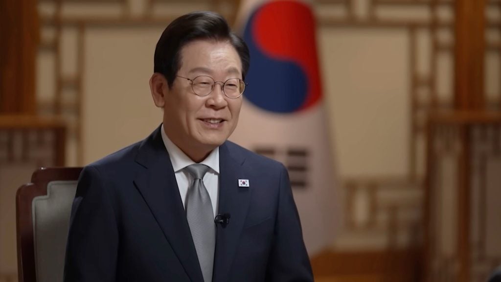 Lee Jae Myung in glasses and a dark suit with a small South Korean flag pin smiles while seated before a blurred South Korean flag and wooden lattice wall.