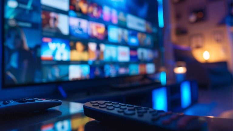 Two remote controls on a glossy coffee table in front of a large TV showing a blurred streaming menu, bathed in blue ambient lighting with a warm lamp glowing in the background.