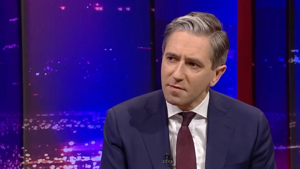 Harris in navy suit and maroon tie with grey hair listening intently on a purple-lit TV studio set.