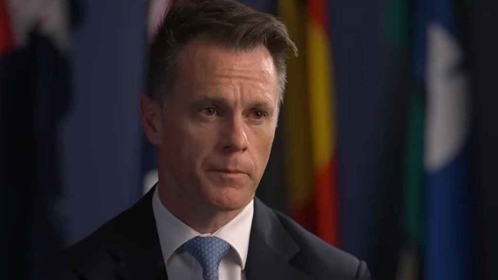 Close-up of a serious middle-aged man in a dark suit and light-blue tie against a blurred row of flags