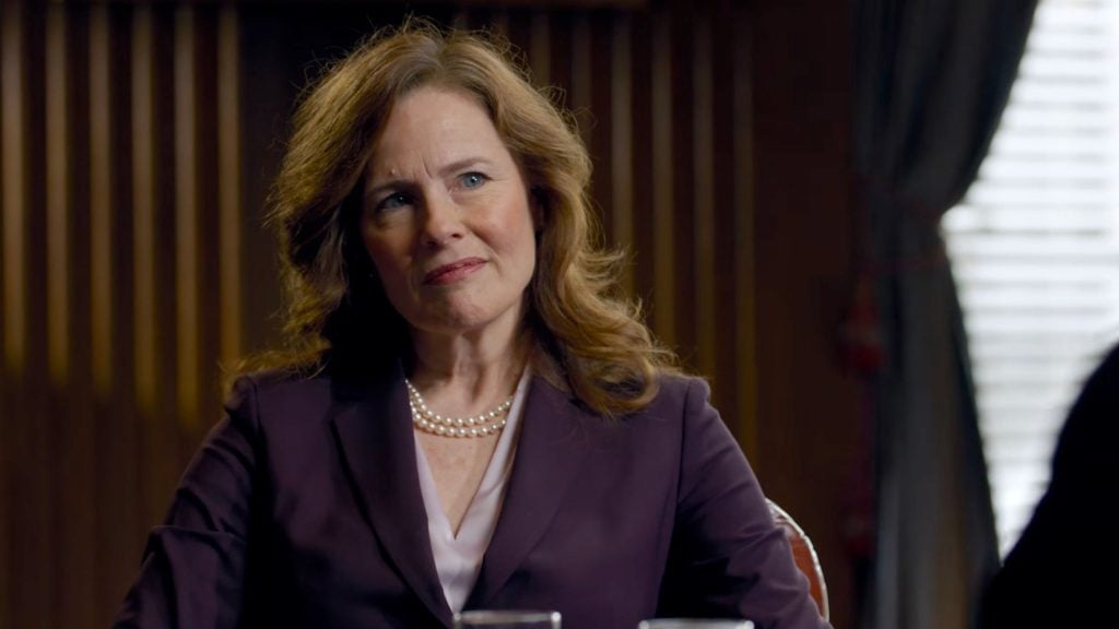 Amy Coney Barrett with wavy brown hair in a purple blazer and double-strand pearls, seated, looking thoughtful.