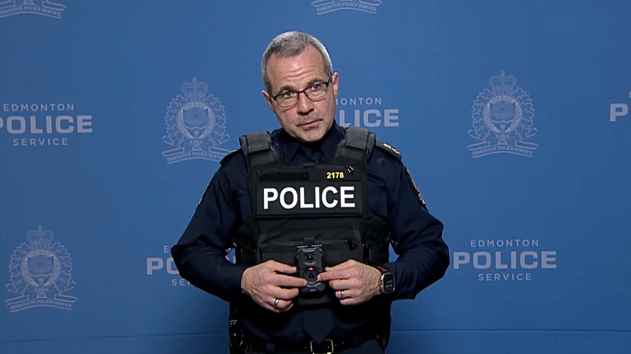 Uniformed police officer wearing glasses and a tactical vest labeled POLICE stands against a blue Edmonton Police Service backdrop, holding a body camera at chest level.