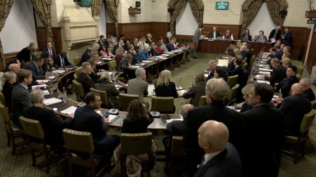 A large formal meeting in an ornate wood-paneled chamber with dozens of MPs seated around U-shaped tables, microphones and papers before them, and officials presiding at a raised bench beneath patterned curtains and wall clocks.