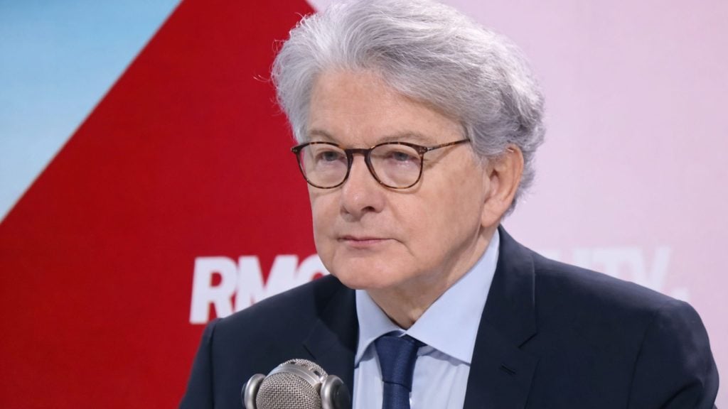 Breton with gray hair and round glasses in a suit speaking into a vintage-style microphone against a red studio backdrop.