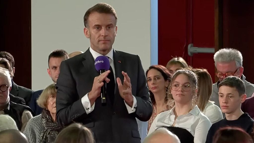 Macron in a dark suit and tie stands at the front holding a purple microphone and speaking to a seated audience, with attentive listeners behind him including a young woman wearing glasses and a teenage boy.