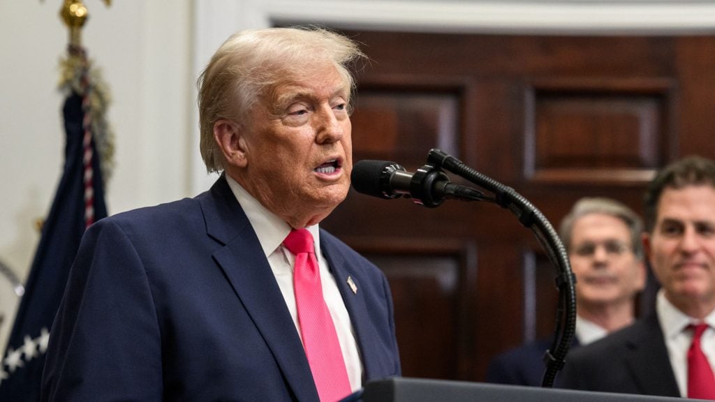Trump in a navy suit and bright pink tie speaking into a microphone at a podium, with two blurred men and a wood-paneled wall in the background.