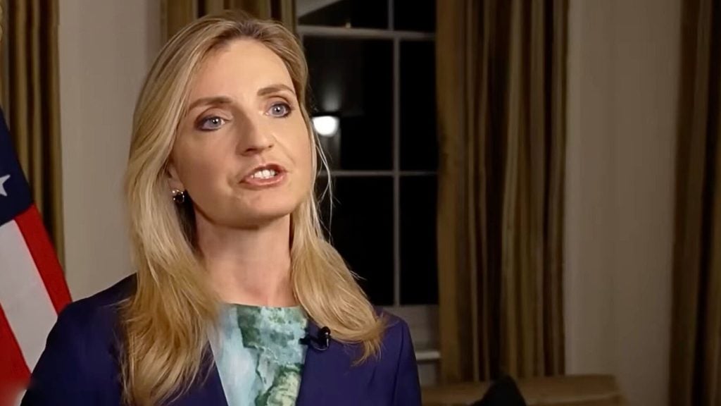 Sarah Rogers speaking in a formal room with an American flag to her left, wearing a blue blazer and green patterned blouse, a window and heavy curtains visible behind her