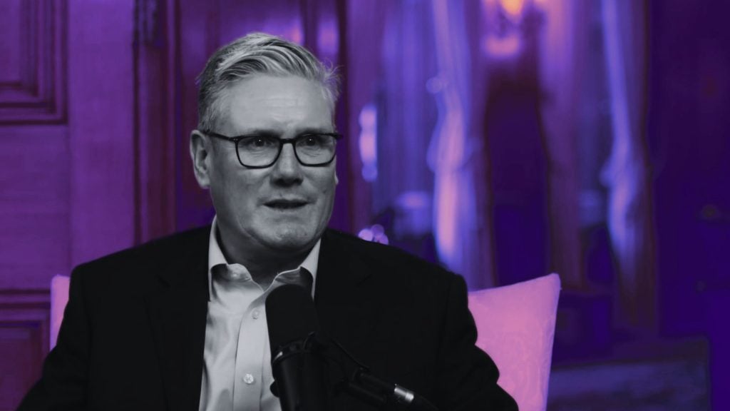 Starmer with gray hair and glasses wearing a dark suit, seated and speaking into a microphone, framed by purple lighting and a blurred curtain and wood-paneled background.