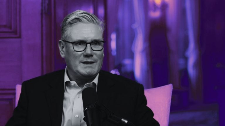 Starmer with gray hair and glasses wearing a dark suit, seated and speaking into a microphone, framed by purple lighting and a blurred curtain and wood-paneled background.