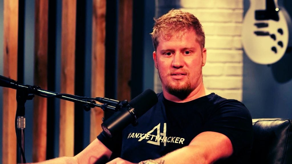 Man with short blond hair and beard speaking into a studio microphone, wearing a black Anxiety Hacker T-shirt.