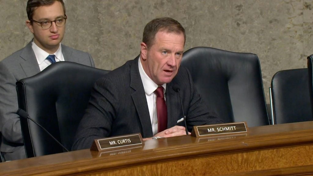 Schmitt in a suit and red tie speaking at a hearing, seated behind a wooden desk with a nameplate and microphone.