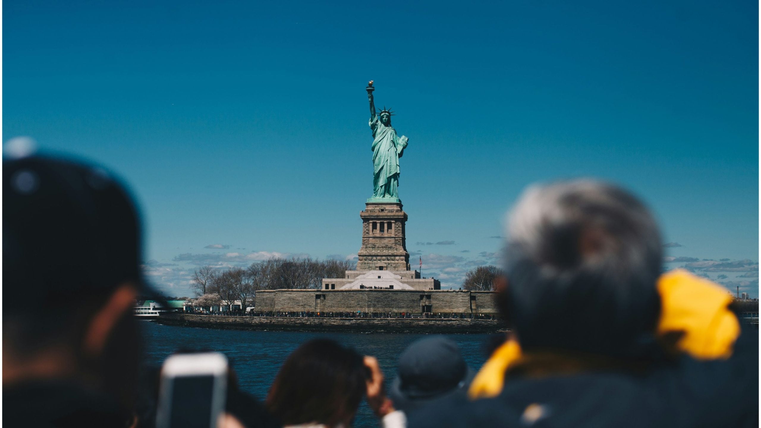 The Statue of Liberty on its stone pedestal on Liberty Island against a clear blue sky, viewed from a boat with blurred tourists in the foreground taking photos.