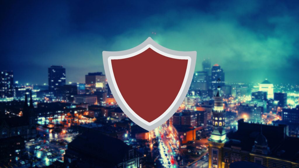 Red shield icon with gray outline centered over a blurred nighttime city skyline with teal sky and glowing street lights.