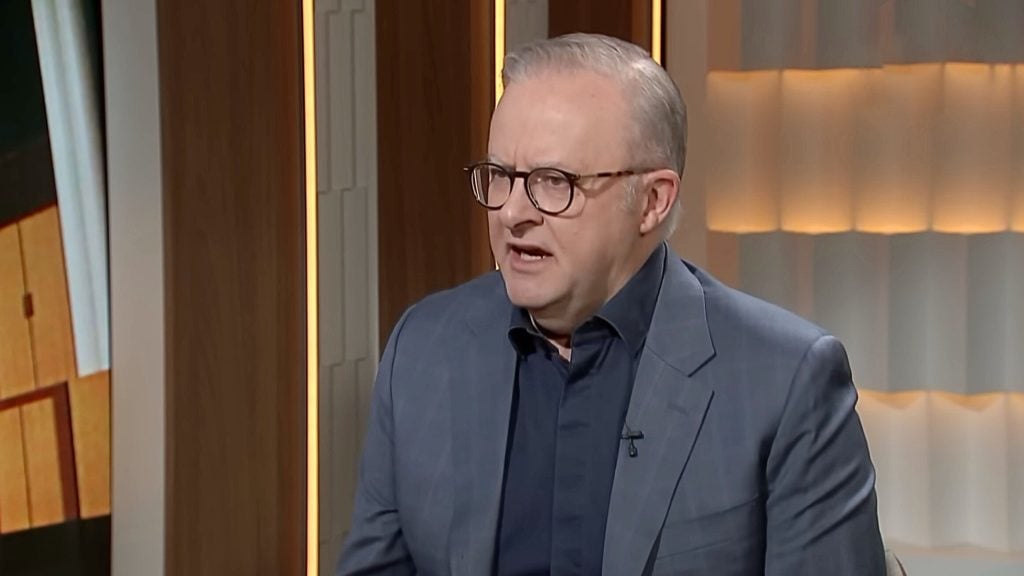 Albanese with short gray hair and round glasses wearing a dark shirt and blue suit jacket speaking on a softly lit studio set with wood paneling and vertical light strips.