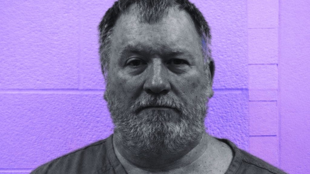 Bearded middle-aged man with short hair and a neutral expression facing the camera against a purple cinderblock wall.